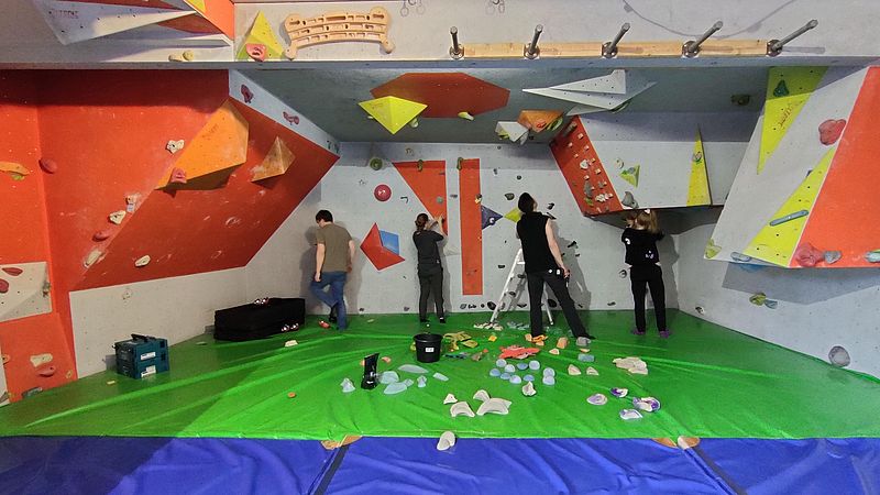 4 Menschen stehen in einer Boulderhalle an der Wand und schrauben Griffe ab. Im Vordergrund liegen viele Griffe auf den Matten.