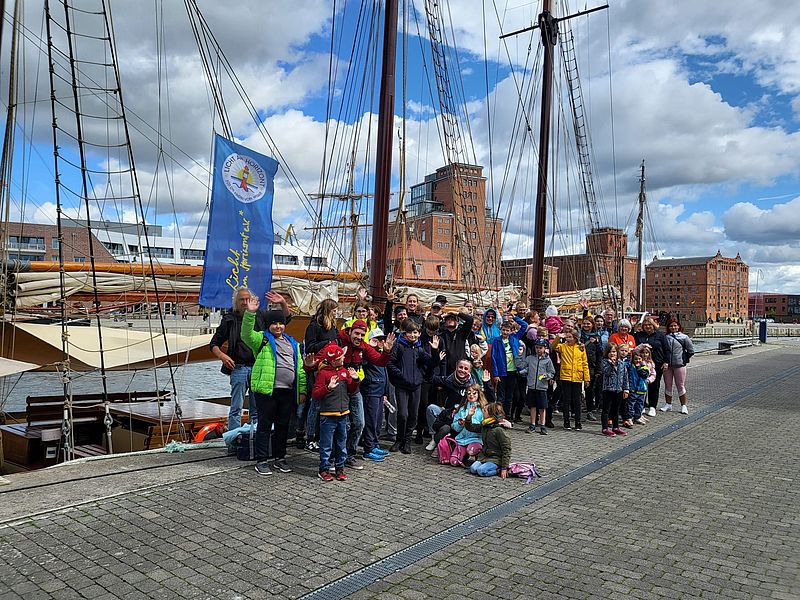 Gruppenfoto von Kindern, Familien und Vereinsmitgliedern am Hafen von Wismar vor einem Segelschiff mit Vereinsfahne.