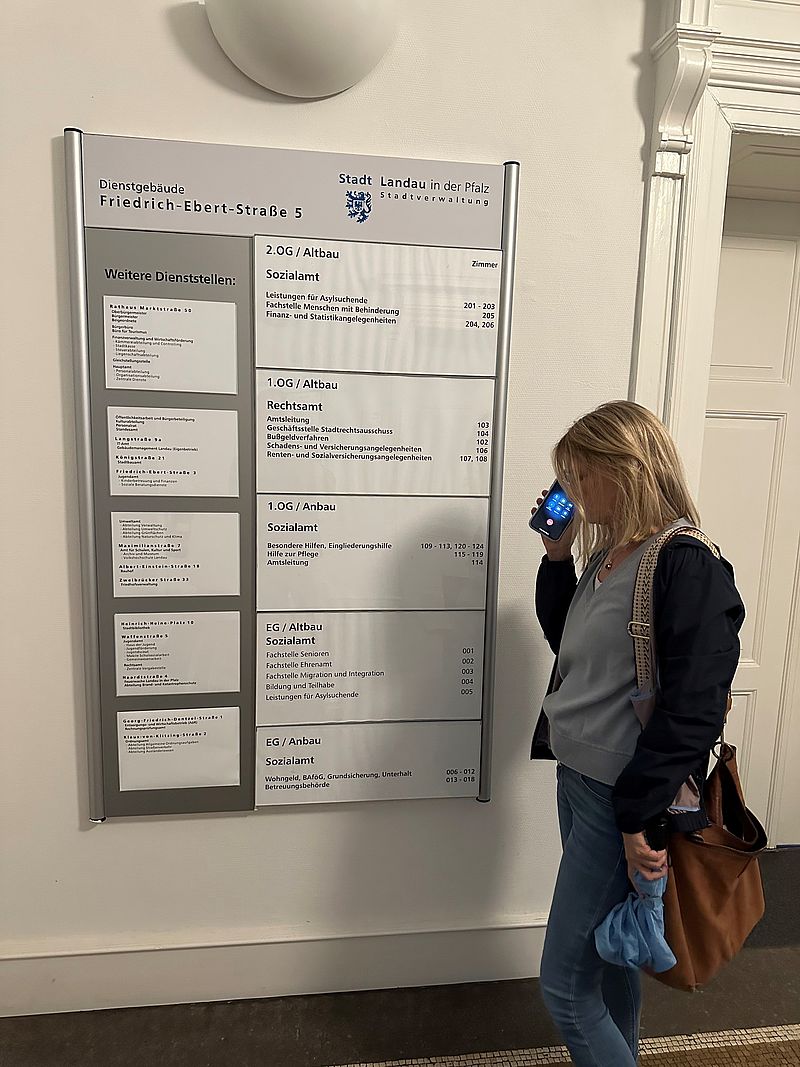 Eine Frau steht mit einem Handy in der Handy vor einem Schild des Dienstgebäudes der Stadt Landau